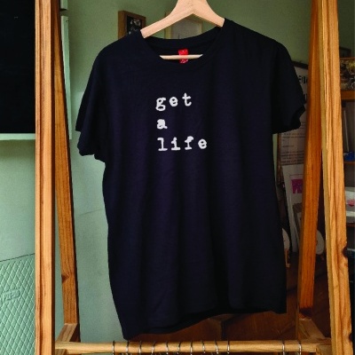 T-shirt preta com texto branco 'get a life' pendurada numa estrutura de madeira