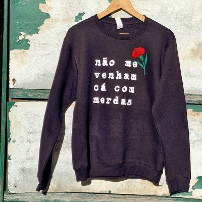 Sweatshirt preta com texto branco e flor vermelha pendurada contra parede verde desgastada.