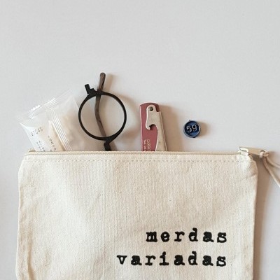 Bolsa de tecido bege com texto 'merdas variadas' e objetos diversos.