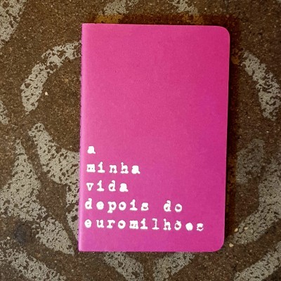 Caderno rosa com texto branco 'a minha vida depois do euromilhões' em chão de pedra