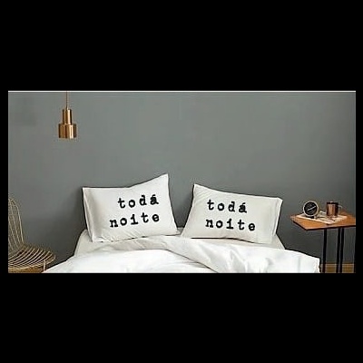 Travesseiros com fronhas escritas, cama branca, parede cinzenta, candeeiro dourado e mesa lateral com objetos