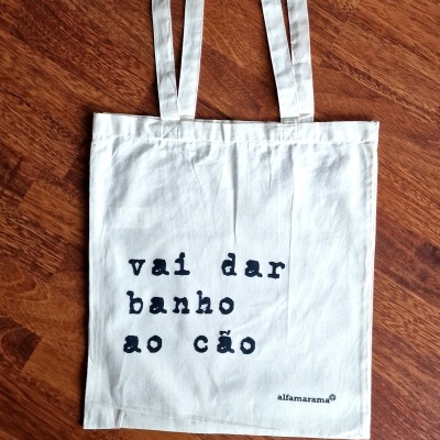 Sacola de tecido branca com texto preto e alças longas sobre chão de madeira