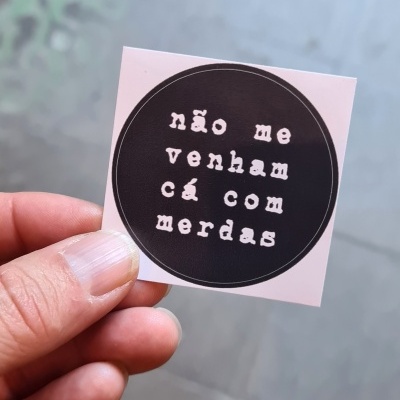 Autocolante quadrado com mensagem em branco sobre fundo preto, segurado por mão, fundo desfocado de chão e grade