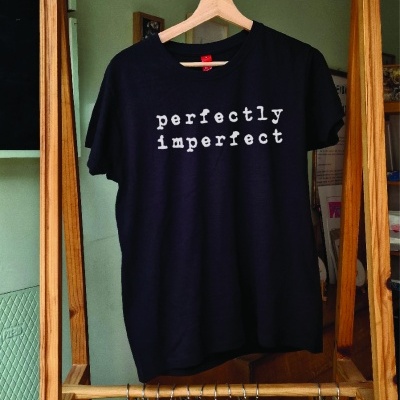 T-shirt preta com texto branco 'perfectly imperfect' pendurada em cabide de madeira