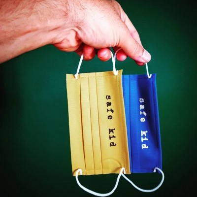 Máscaras faciais amarela e azul com texto 'safe kid' penduradas num dedo