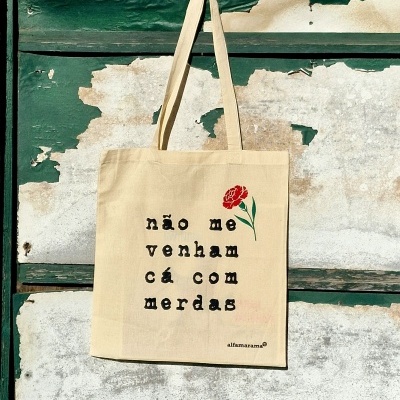 Saco tote bege com texto e flor pendurado numa porta verde antiga