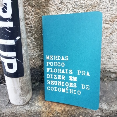 Livro ou caderno azul turquesa com texto branco encostado no chão junto a poste e parede de pedra