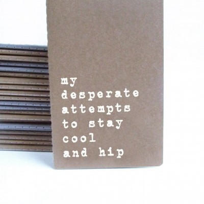 Caderno castanho claro com texto branco 'my desperate attempts to stay cool and hip'
