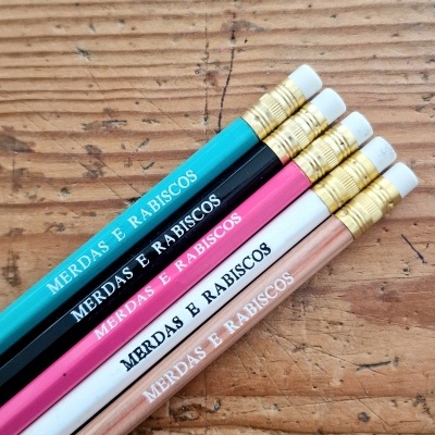 Cinco lápis coloridos com texto 'MERDAS E RABISCOS' numa mesa de madeira