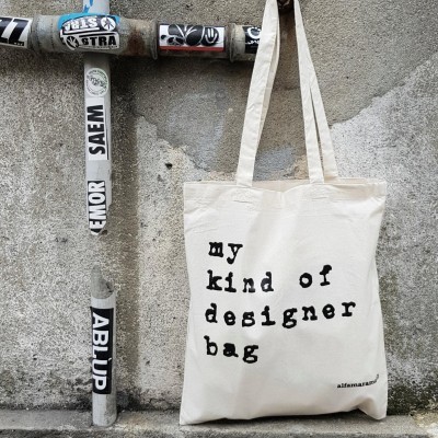 Saco de tecido bege com texto impresso preto pendurado numa parede cinzenta ao lado de tubos com autocolantes