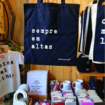 Saco de pano azul com texto branco pendurado, rodeado por artigos de papelaria e merchandising em bancada de madeira