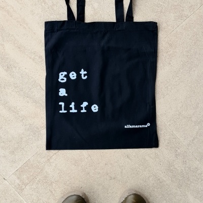 Saco de tecido preto com texto branco get a life colocado no chão junto a sapatos castanhos e calças verdes visto de cima