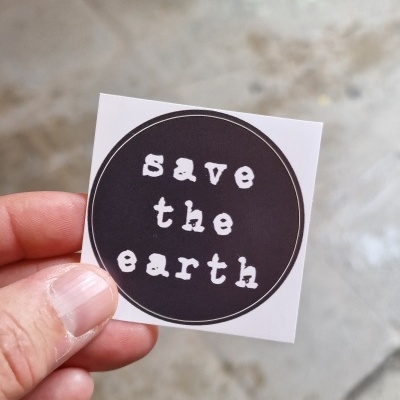 Etiqueta quadrada branca com círculo preto e texto 'save the earth' segurada por uma mão