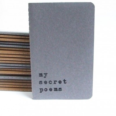 Caderno cinzento com texto 'my secret poems' e cadernos castanhos ao fundo