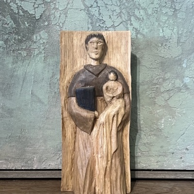 Escultura em madeira de duas figuras, uma masculina com livro azul e uma feminina, contra parede verde texturada.