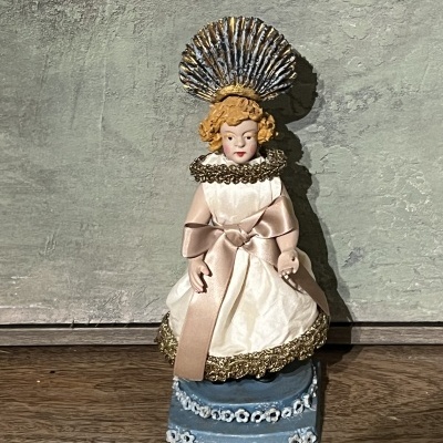 Boneca decorativa com vestido branco e adorno prateado na cabeça sobre base azul com flores.