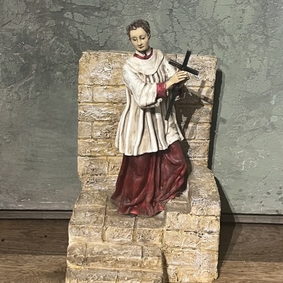 Estátua decorativa de jovem com traje litúrgico branco e vermelho sentado em degraus de pedra.