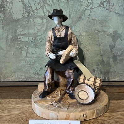Escultura de figura humana em madeira com chapéu, avental e ferramentas, sobre base redonda de madeira
