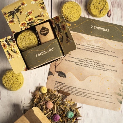 Caixa floral aberta com discos amarelos, pacote de papel com logo, cartão com texto em espanhol e ervas coloridas espalhadas.