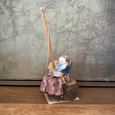 Figura decorativa de pessoa sentada em tronco a segurar pau comprido, madeira pintada, fundo cinzento texturizado