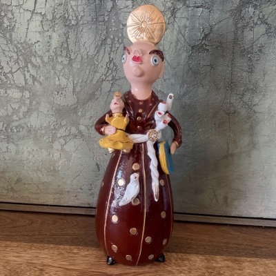 Figura cerâmica pintada de personagem com vestido castanho e pontos dourados segurando boneca amarela e pássaros brancos