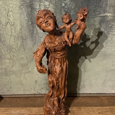 Estátua de cerâmica castanha com figura feminina e bebé, sobre madeira, contra parede texturada.
