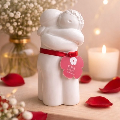 Estatueta branca de duas figuras a abraçar com fita vermelha e etiqueta rosa, flores e vela ao fundo