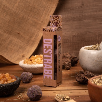 Embalagem de incenso Destrabe em papel reciclado com padrão geométrico, rodeada por resinas, ervas secas e objetos naturais sobre mesa de madeira