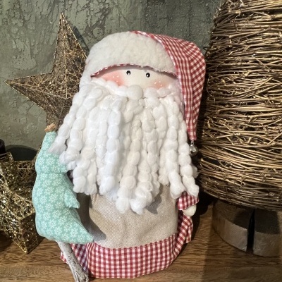 Boneco de Natal em tecido com barba branca e chapéu xadrez, segurando árvore azul com decoração à volta