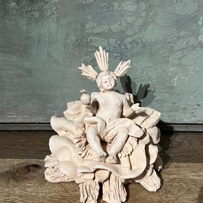 Figura escultórica bege de bebé num trono decorado sobre madeira e fundo cinza esverdeado