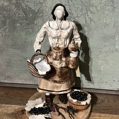 Estatueta de mulher tradicional com cesta e saco e base decorada com uvas e folhas
