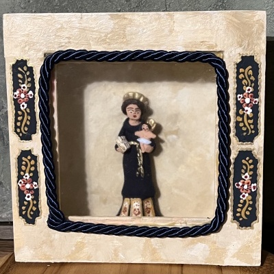 Quadro decorativo com moldura creme e figura de santo em tecido segurando criança