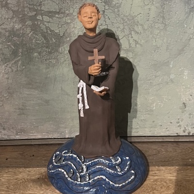 Figurinha de frade de túnica castanha segurando cruz e livro sobre base azul com padrões ondulados.