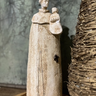 Estátua em madeira clara de figura religiosa com criança ao lado de objeto de ramos entrelaçados