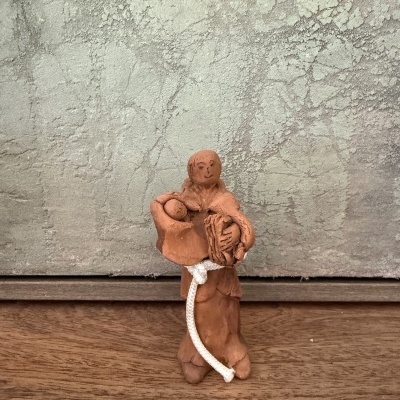 Estátua de barro rústica com figura humana e cordão branco sobre madeira