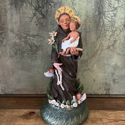 Estátua religiosa de santo em resina com criança, flores e anjos, base verde com relevo, sobre superfície de madeira.