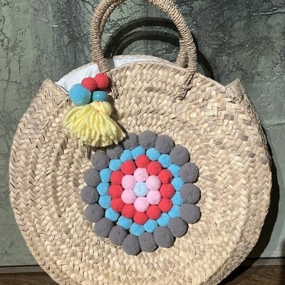 Mala redonda de palha com pompons coloridos e tassel pendurado.