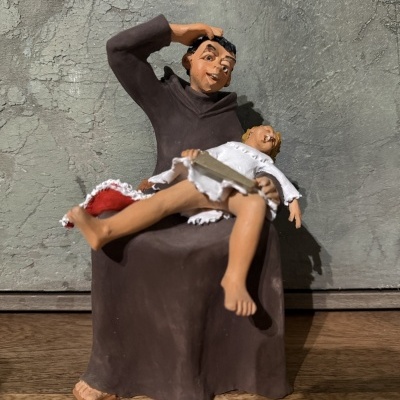 Estatueta de duas pessoas, uma sentada com túnica marrom e outra deitada com vestido branco, em fundo cinzento e superfície de madeira.