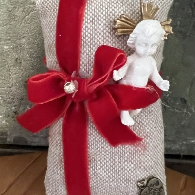 Almofada pequena bege com laço vermelho, anjo branco e detalhe metálico em forma de coração