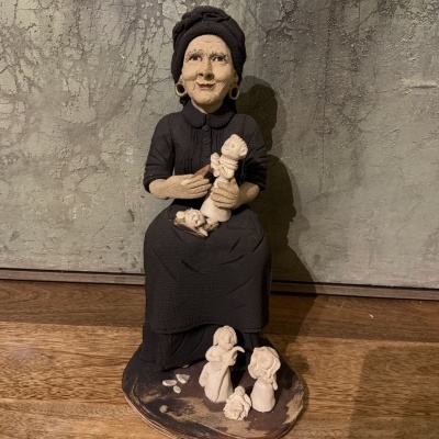 Estátua de senhora sentada com vestido preto segurando figuras em cerâmica sobre base com figuras pequenas adicionais