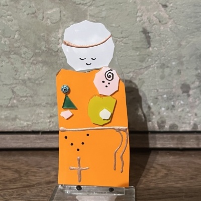 Objeto decorativo em madeira com corpo laranja e cabeça branca com rosto desenhado simples.