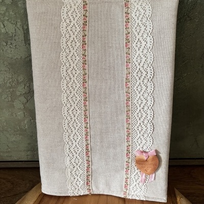 Capa de tecido bege decorada com renda branca e fitas floridas, com pendente de madeira em forma de coração