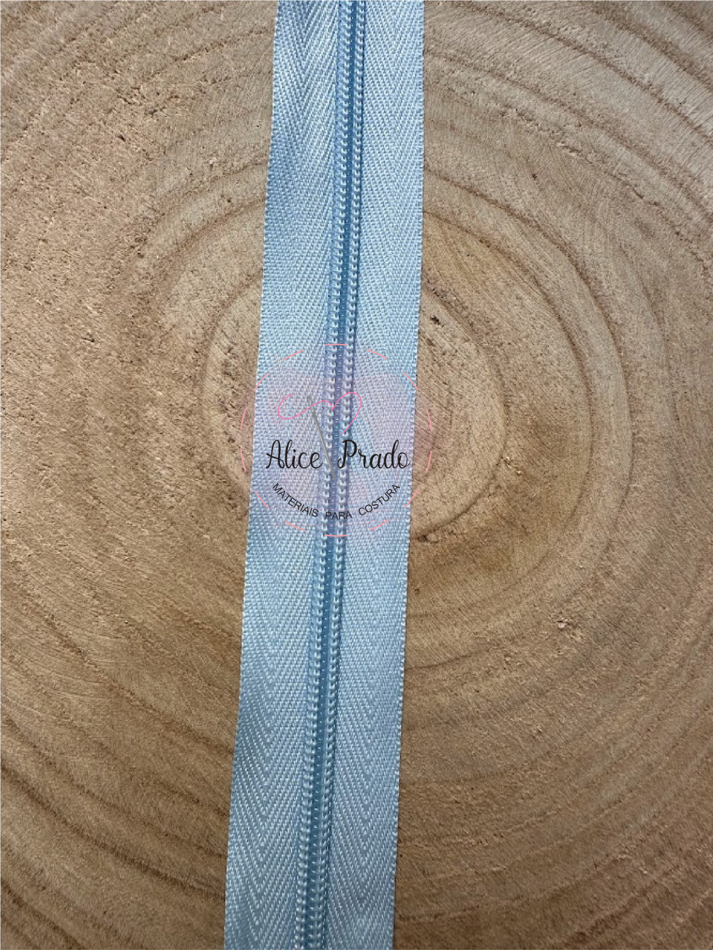 Fecho éclair azul claro sobre superfície de madeira com etiqueta 'Alice Prado'.