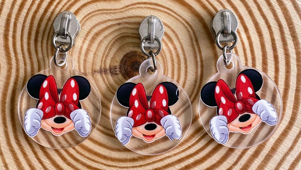 Três chaveiros redondos com Minnie Mouse a tapar os olhos, laço vermelho com bolinhas, sobre superfície de madeira circular.
