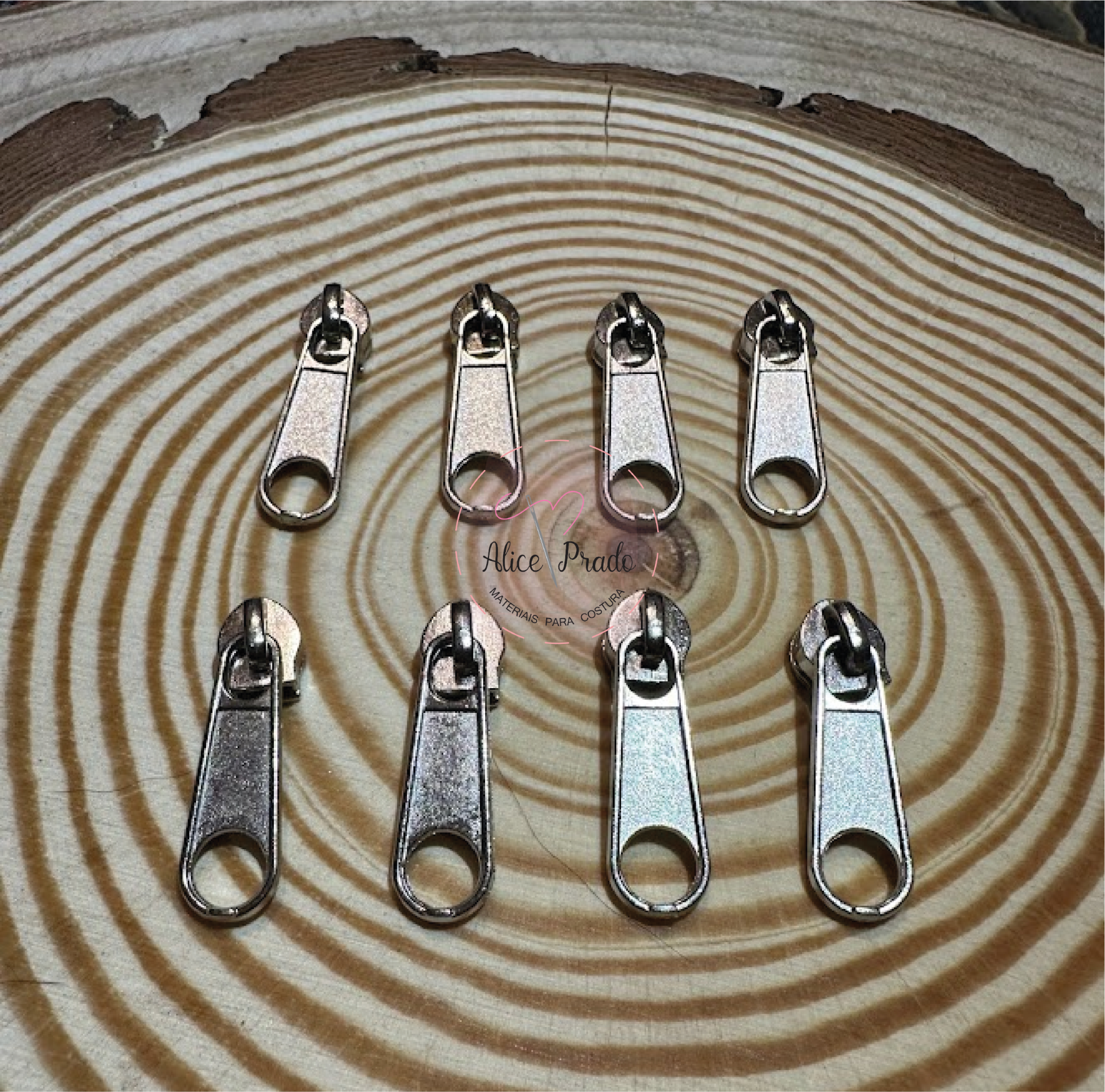 Cursoras metálicas prateadas para fechos éclair sobre madeira com padrão circular e texto 'Alice Prate'