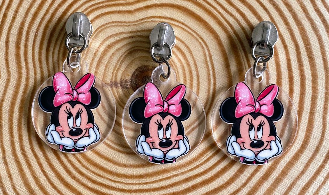 Três chaveiros redondos transparentes com a personagem Minnie Mouse em laços rosas sobre fundo de madeira