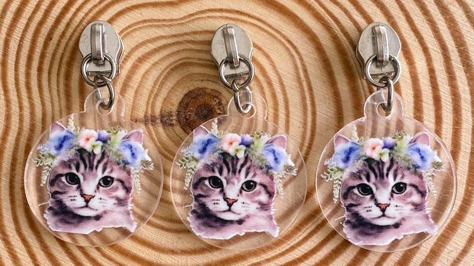 Três chaveiros de gato com flores, pendurados em prendedores metálicos, sobre madeira