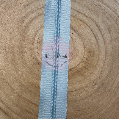 Fecho éclair azul claro sobre superfície de madeira com etiqueta 'Alice Prado'.