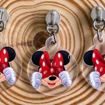 Três chaveiros redondos com Minnie Mouse a tapar os olhos, laço vermelho com bolinhas, sobre superfície de madeira circular.