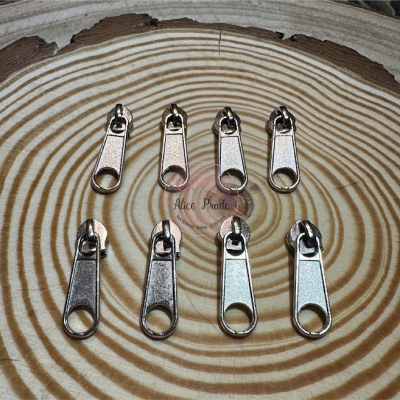 Cursoras metálicas prateadas para fechos éclair sobre madeira com padrão circular e texto 'Alice Prate'